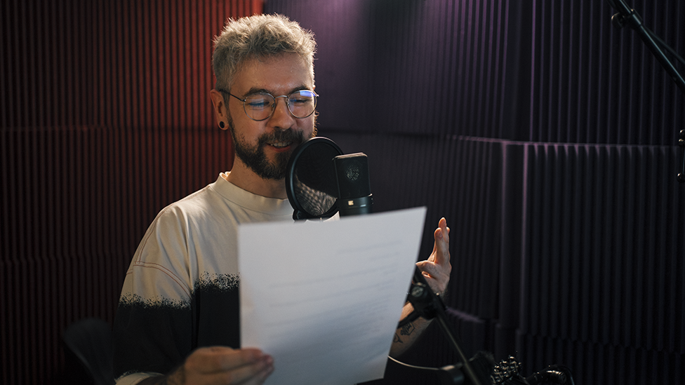 Jacksepticeye stands next to a microphone in a recording booth with red, ridged soundproof walls. He's holding a piece of paper in one hand, gesturing with the other, as he reads from the script.