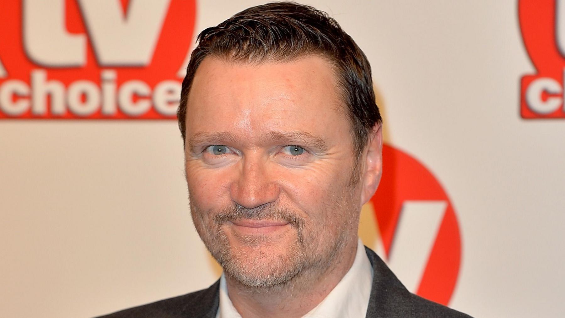 Ian Puleston-Davies is wearing a suit and smiling at the camera. He has dark hair and beard