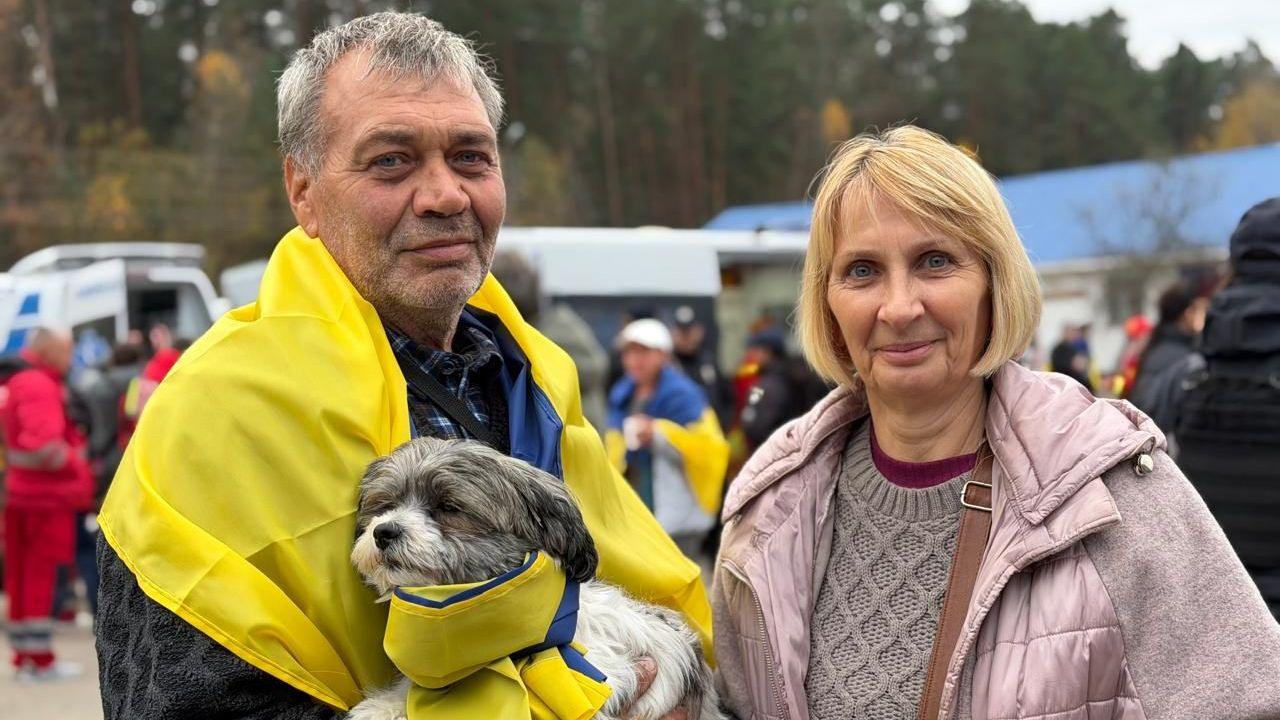 Viacheslav, who has grey hair and is draped in a Ukrainian flag, holds a small dog in his arms. He is stood next to his wife Olha, who has a blonde bob. Both are looking at the camera.
