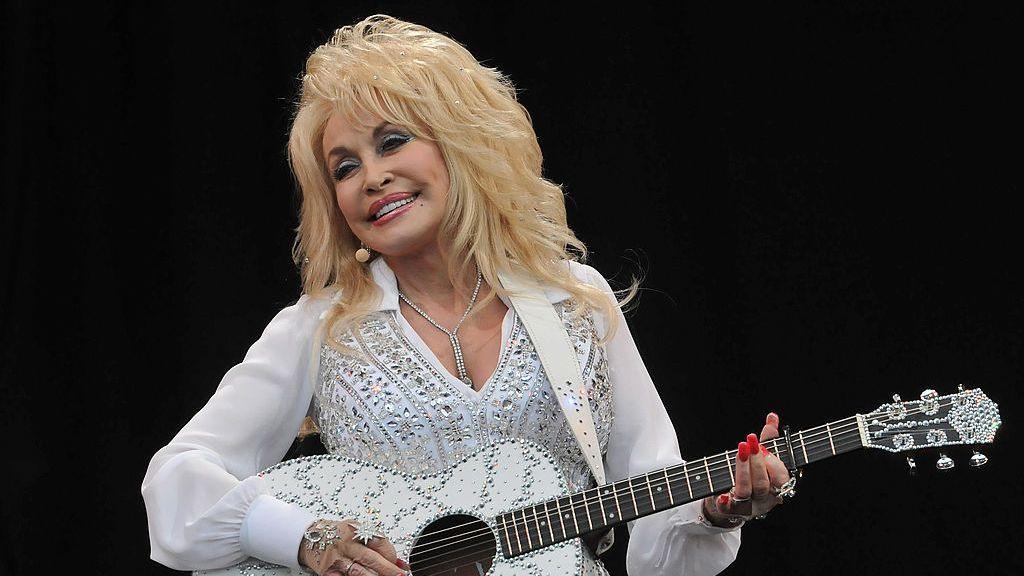 Dolly Parton on stage at Glastonbury. She is wearing a rhinestone-encrusted all-white outfit and holds a matching rhinestone-encrusted guitar