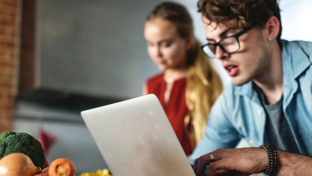 How the Budget affects you Man looks at laptop with a woman cooking behind him and some vegetables on a work surface behind the laptop.