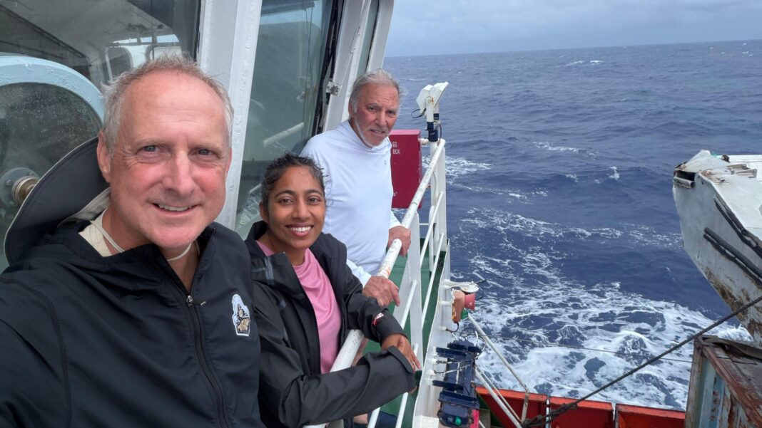 Two men and a woman stand on the viewing deck of a vessel travelling through the Pacific Ocean.