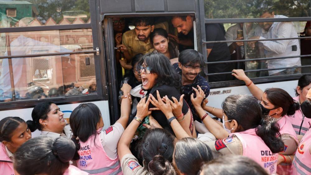 A bus packed with people as a woman and man are being pushed on to the vehicle by people in pink uniforms.