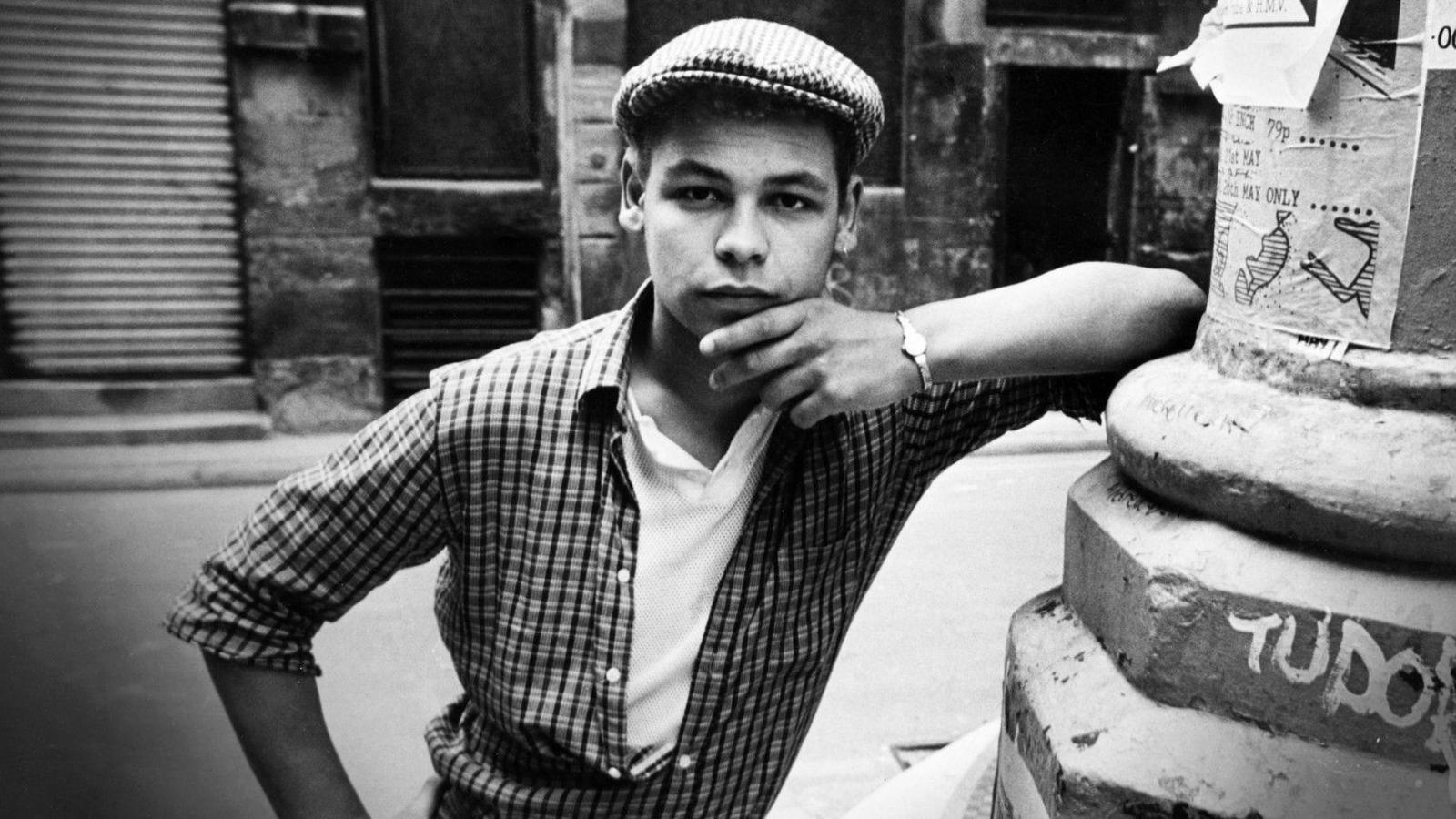 A black and white photograph of Craig Charles wearing a checked shirt over a t-shirt and a flat cap. He is leaning against a pillar in.