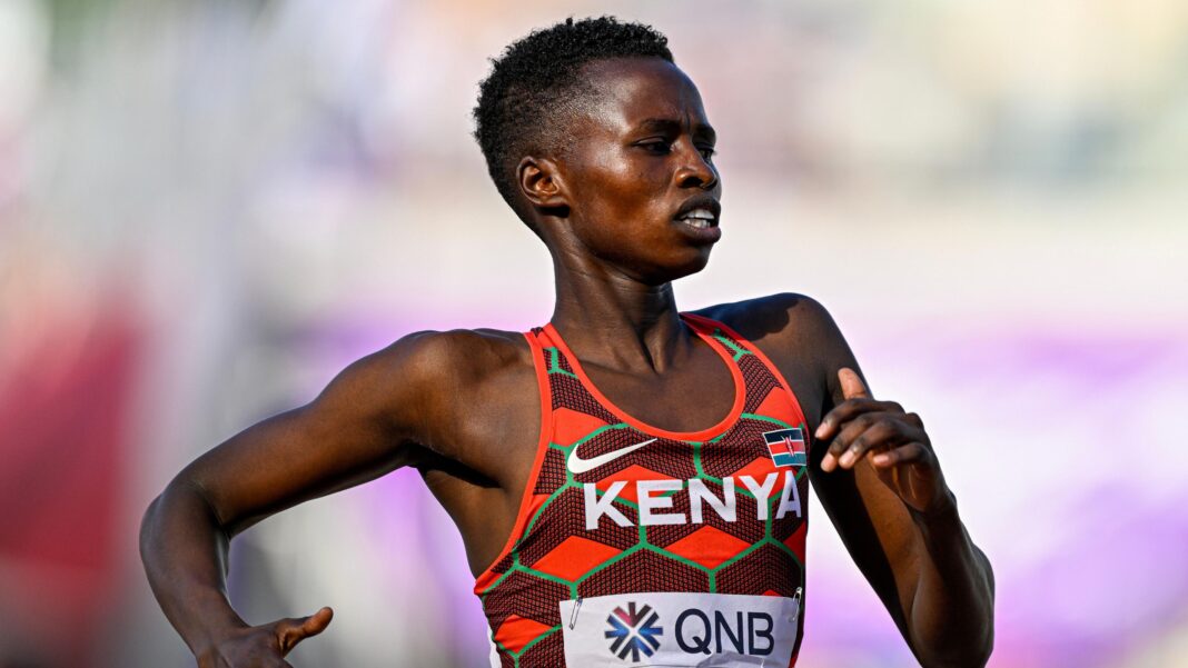 Naomi Korir pictured from stomach up running from left to right during a race. She wears a Kenya running vest made of up light and dark red diamond and chevron patterns with thin green grid lines running over the top and the word 'Kenya' in white letters across the chest. Korir has a fixed expression exuding effort and is pumping her arms hard