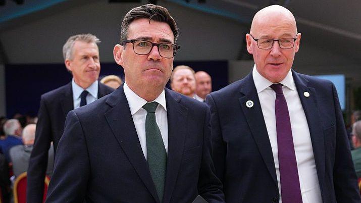 Andy Burnham has black hair and is wearing black glasses, a blue jacket, white shirt and green tie as he stands beside John Swinney- a bald man wearing black glasses, a blue shirt and purple tie