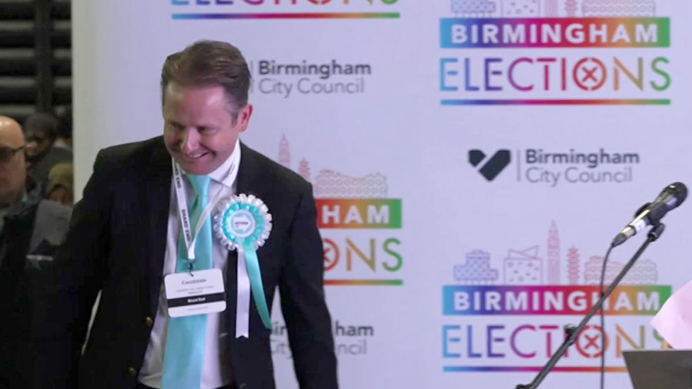 Alan Feeney on the stage after being elected a councillor at election in May 2026. He is wearing a dark jacket and white shirt with a light blue Reform coloured tie and a similar coloured rosette. There is election branding behind him.