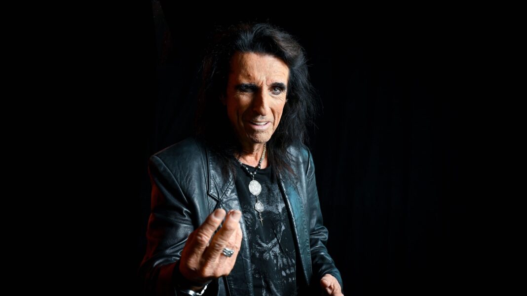 ‘Alice Cooper is a character, like Jekyll and Hyde’ A studio-style portrait shows a person standing against a black backdrop, wearing a black leather jacket over a dark graphic T-shirt and several necklaces. The person has long dark hair and extends one hand towards the camera, creating a sense of depth in the image.