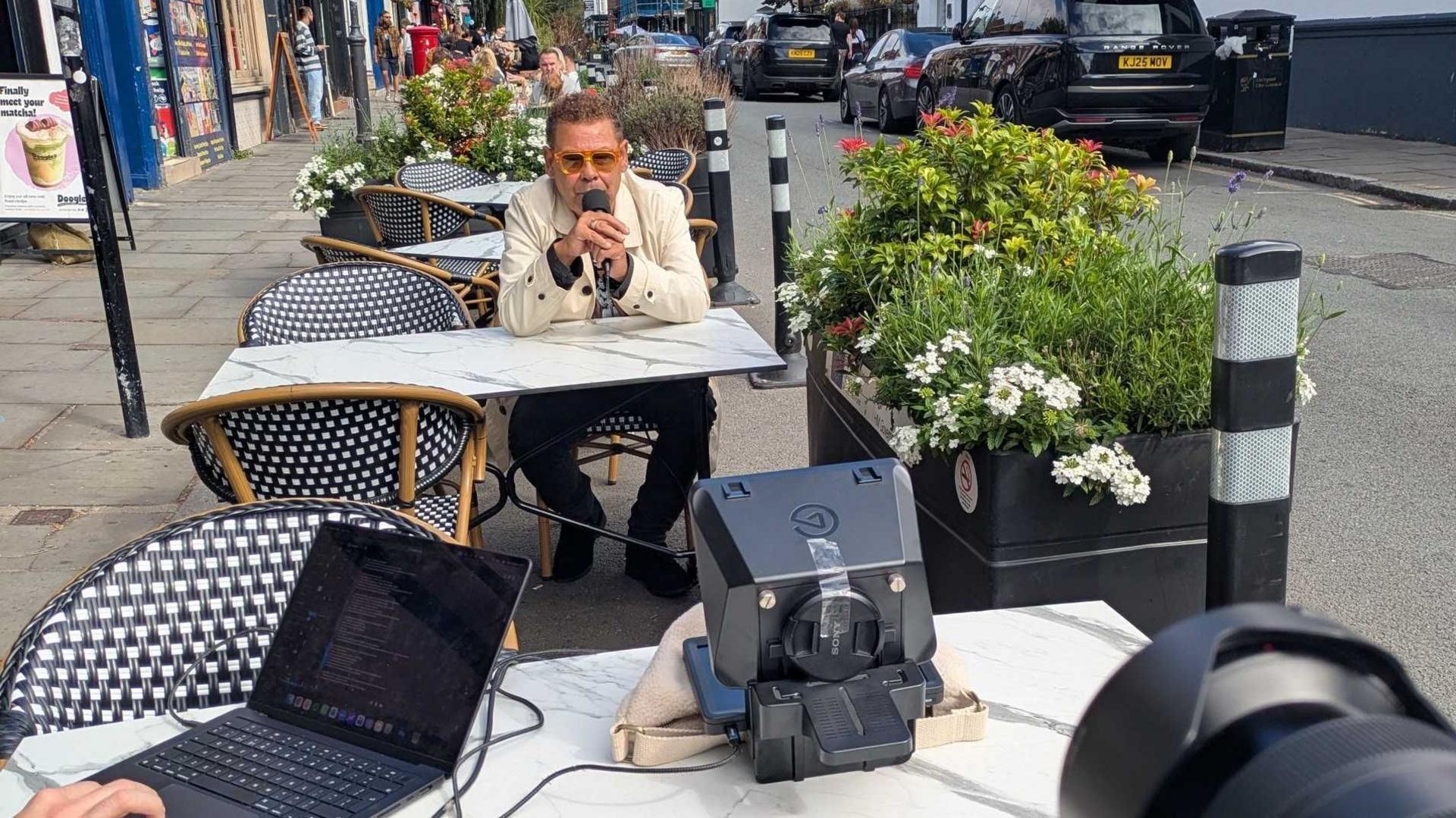 Craig Charles is sat at a table outside a cafe in Liverpool speaking into a microphone as he is filmed for his spoken worked film.