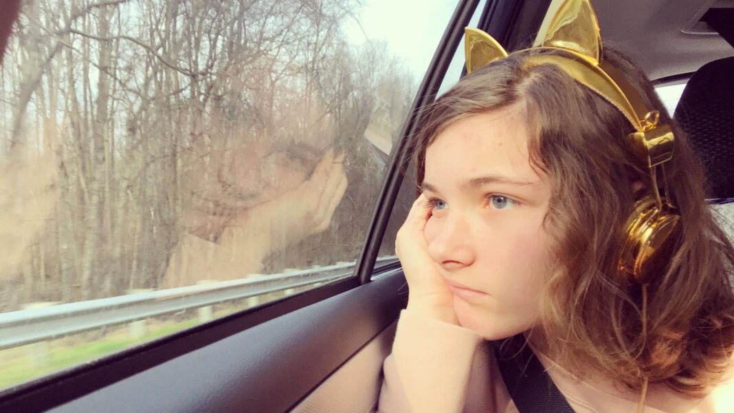 EV drivers love their cars, but some passengers feel sick – this could be why A girl sits in the backseat of a car. She is resting her head in her hands and is wearing gold headphones with animal ears. She looks unhappy or bored.