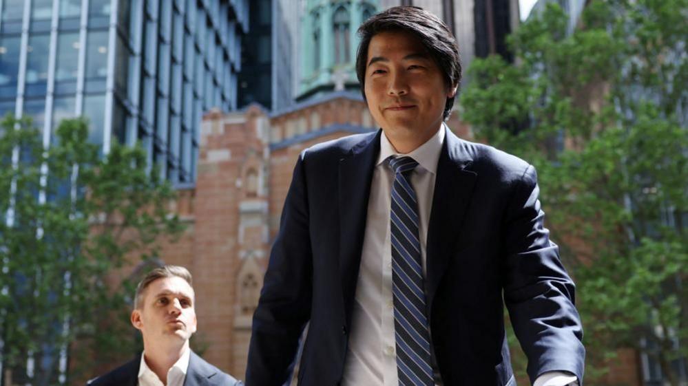 A man with short dark hair wearing a suit walks on a street with buildings and trees in the background