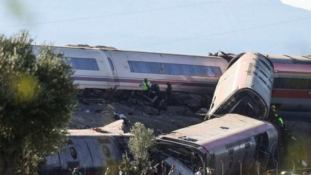 Three overturned train carriages lying on an embankment, with others from another train tilted over. Emergency workers can be seen helping people