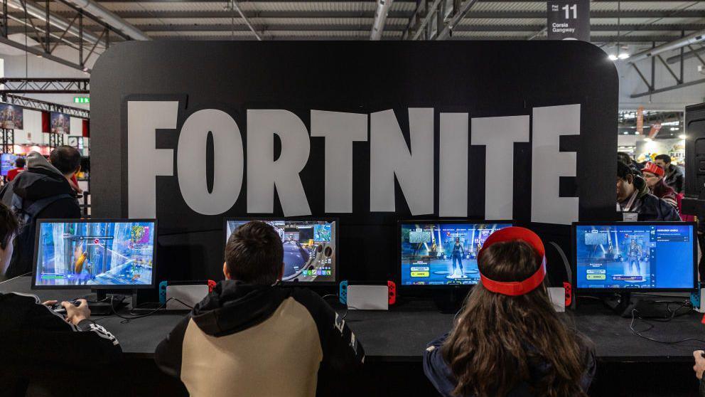 A group of children playing Fortnite on computers at a gaming exhibition, with the game's logo emblazoned on a sign above them.