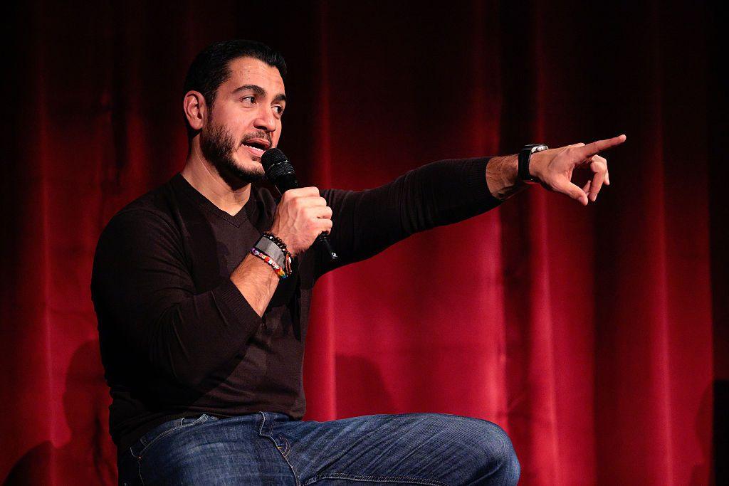 Abdul El-Sayed sits on a stage in front of a red curtain, wearing a black V-neck, long-sleeved T-shirt and blue jeans. Holding a microphone in one hand, he gestures with the other while speaking.