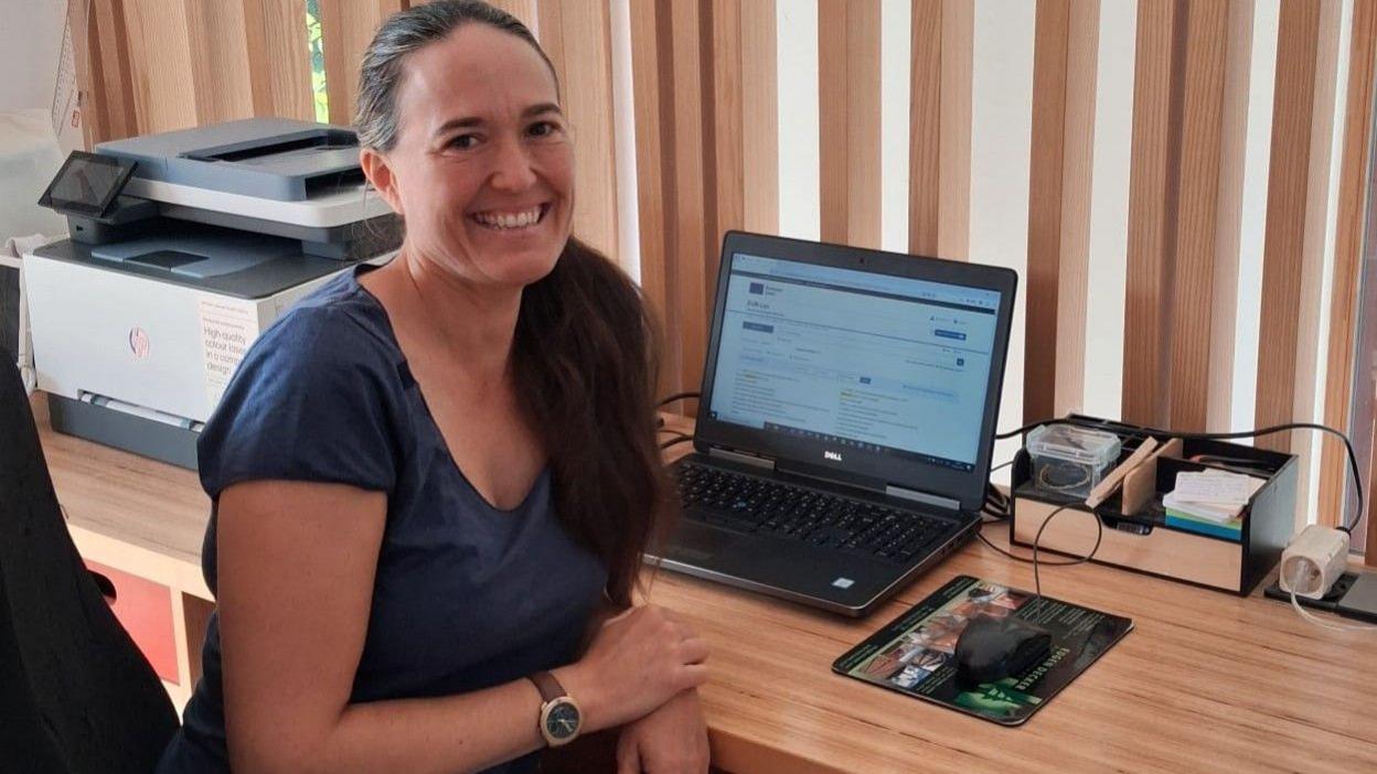 Smiling and with long brown hair, Ariella Baltié sits in front of her laptop