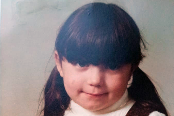 An old portrait picture of Tracy Lewis as a young girl. She has dark hair tied in pigtails and a long fringe, and is wearing a white top and a brown dress.