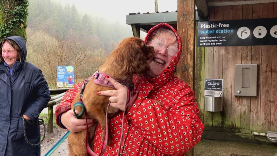Wet looking woman in red coat holds dog