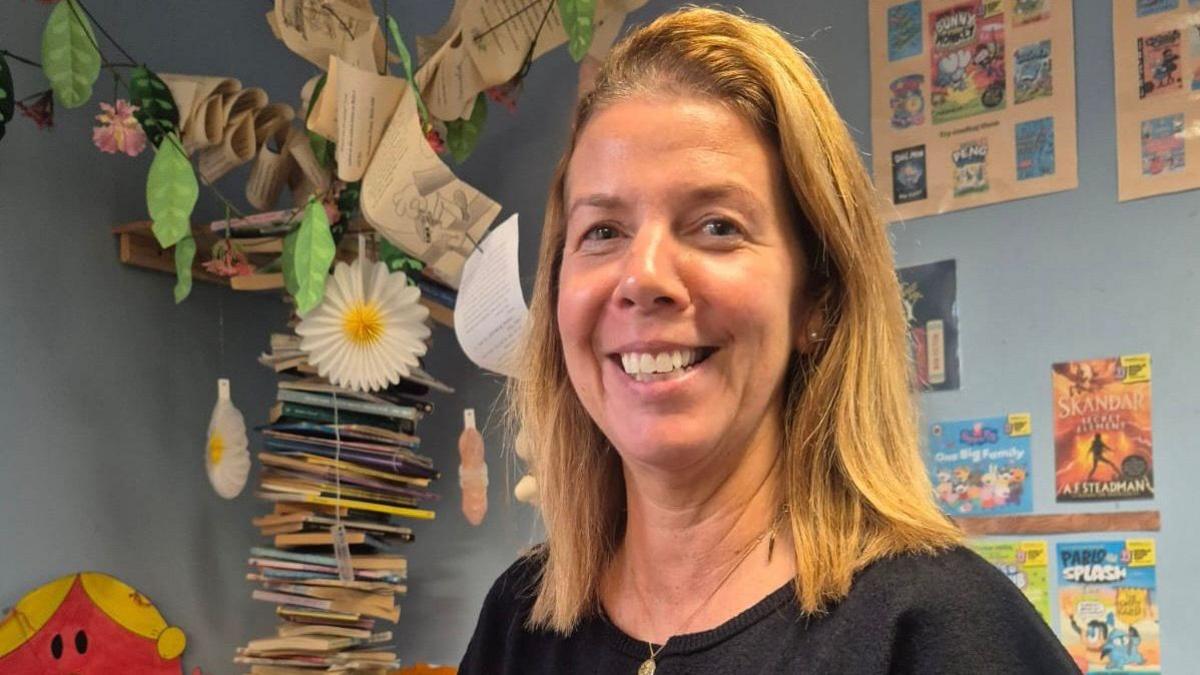 Kirsty Logan-Hall - a woman with shoulder-length blonde hair - smiles at the camera in a classroom setting.  She is wearing a black top and a gold pendant necklace.