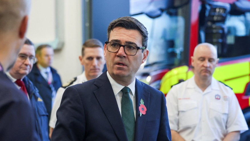 Head and shoulders shot of Burnham meeting firefighters, who are visible in the background along with a fire engine. He is wearing a navy suit, a white shirt and a green tie, with a poppy on his lapel.