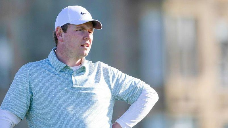 Robert MacIntyre stands as he wears light-coloured polo shirt, with white long-sleeved undershirt, and white golf cap
