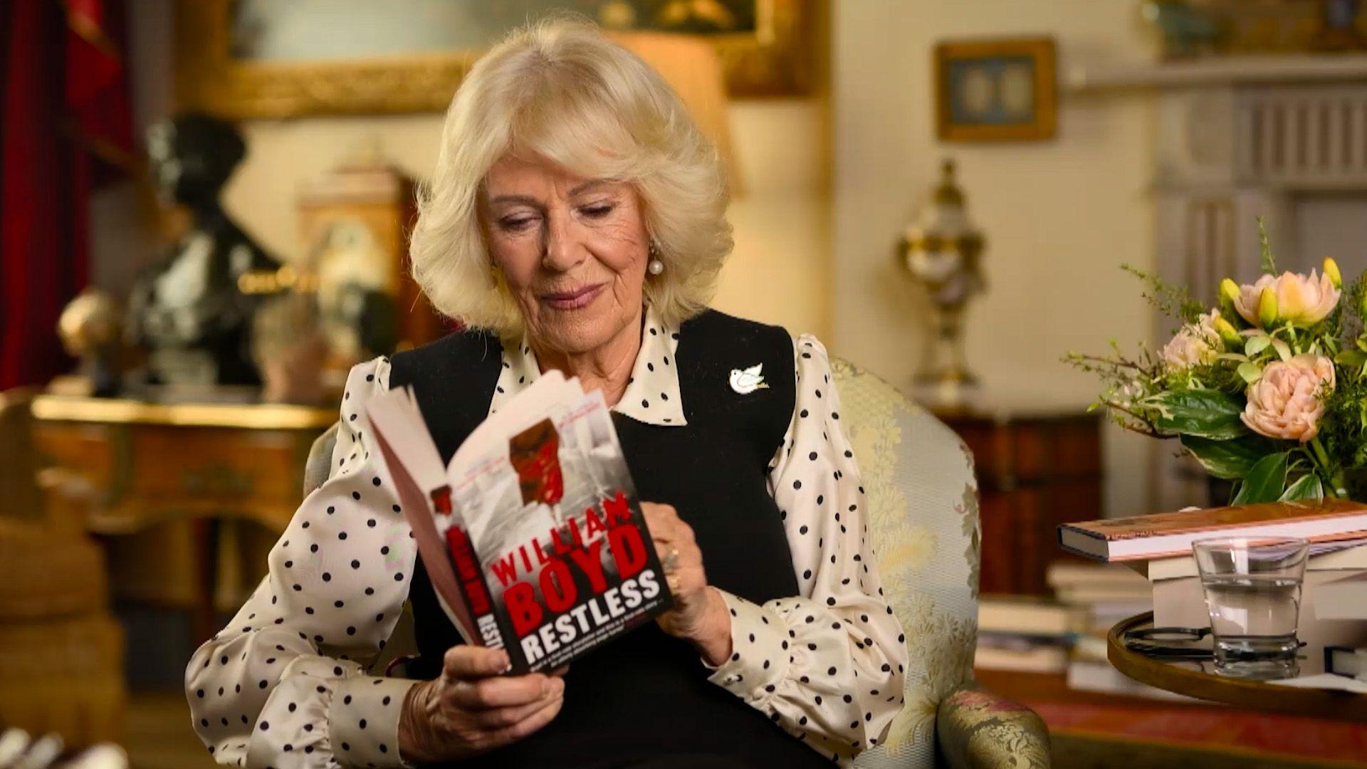Queen Camilla sits in a chair reading a copy of William Boyd's Restless