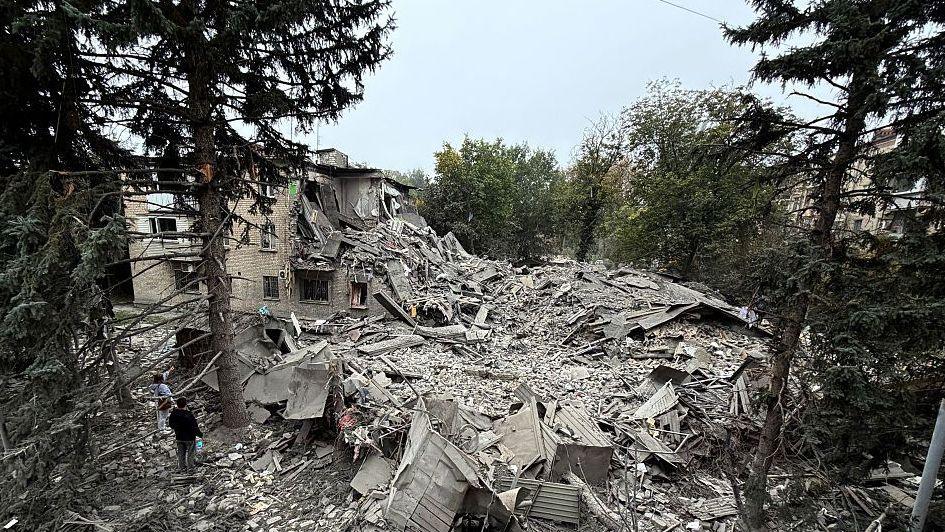 A wide angle photo of an apartment building that was hit by a Russian glide bomb. Most of the structure is completely flattened. Trees surrounding the site are damaged and covered in dust.