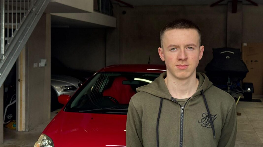 A young man in a green hooded jacket stands in front of a red car which is parked in a garage
