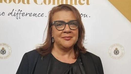 Andri Nicolau, a woman with dark hair, glasses and a dark jacket is stood in front of a colourful wall at an awards ceremony.