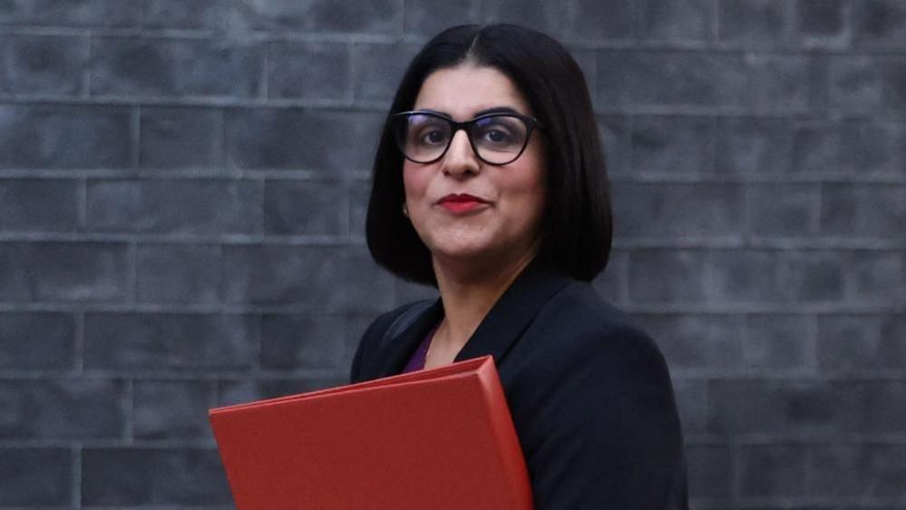 Home Secretary Shabana Mahmood carries a red folder as she walks past a grey brick wall.