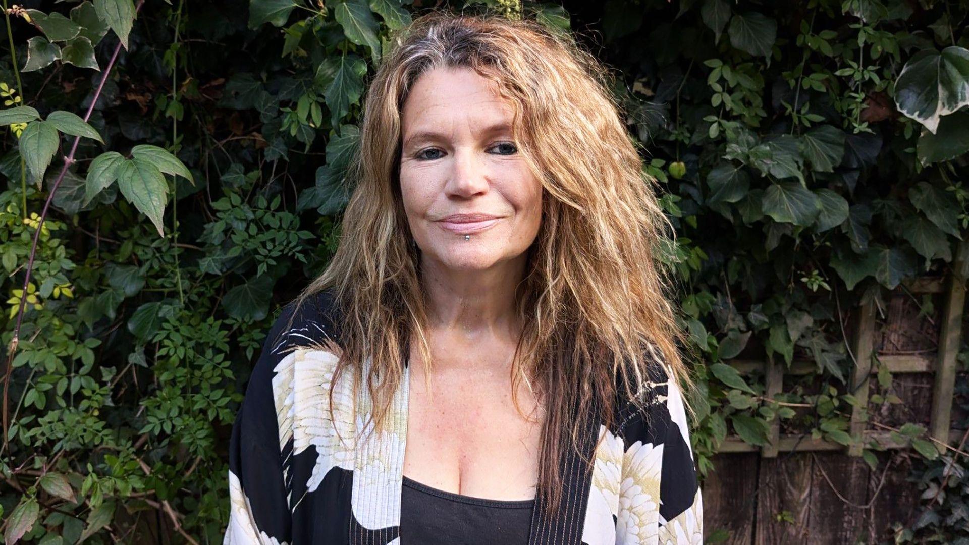 Helen is standing in front of a garden fence. She has long hair and is wearing a black top with a black and white cardigan over it