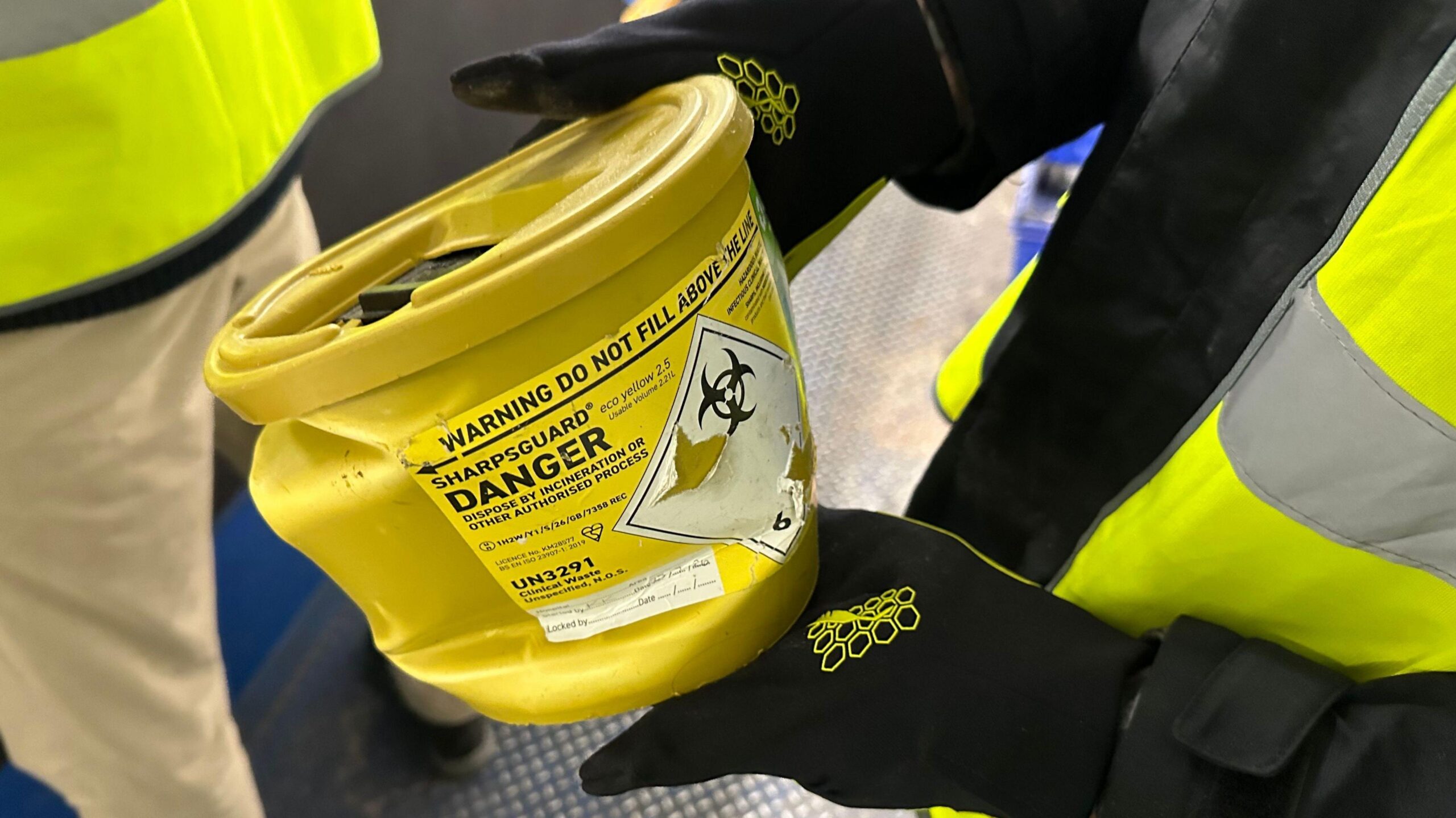 Close-up of a worker wearing black protective gloves and a high-visibility jacket holding a yellow clinical sharps container inside a recycling facility. The container bears a biohazard symbol and warning labels instructing users not to fill it above the marked line.