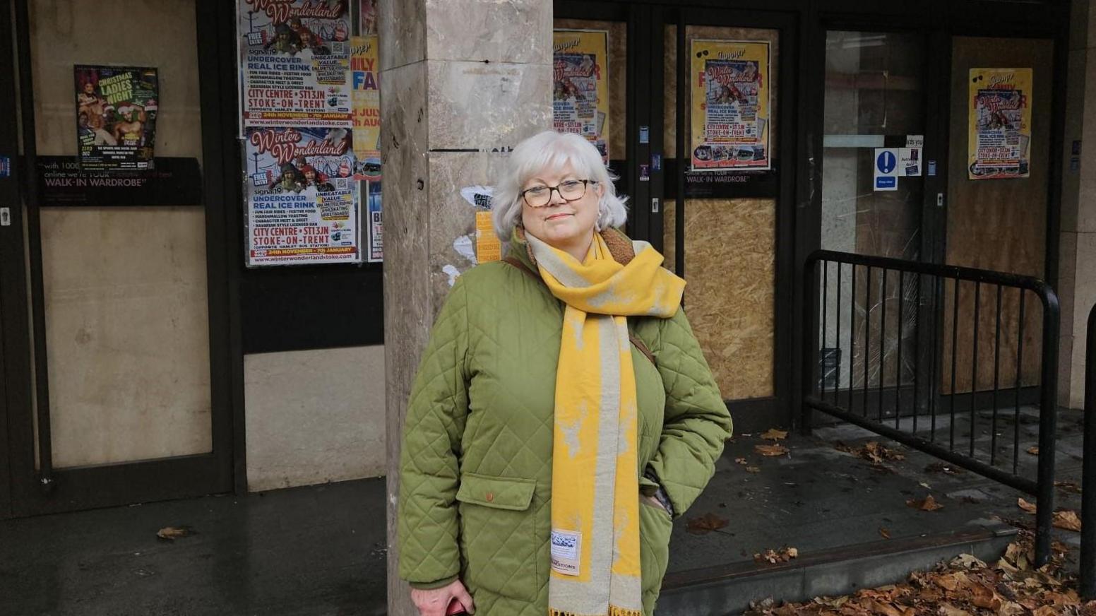 Jane Gynn, a woman with short silver hair, wears a green quilted coat and yellow and grey scarf. She stands outside the doors of a closed building, all of the doors are missing glass and have been boarded up and covered in posters