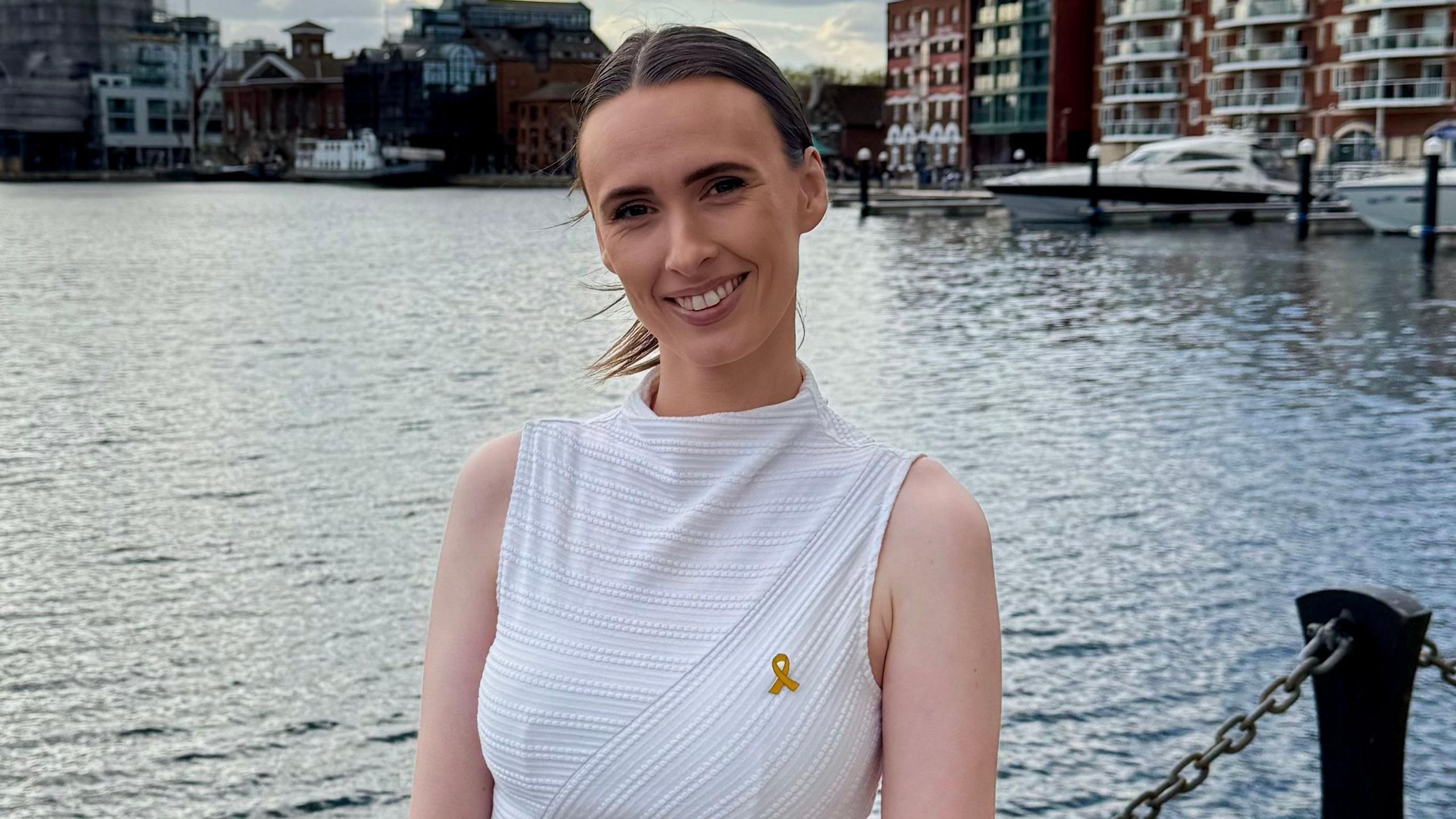 A woman with brown hair that is tied up in a ponytail smiling, she is wearing a white sleeveless top and has a breast cancer awareness pin on, the background is a dock.