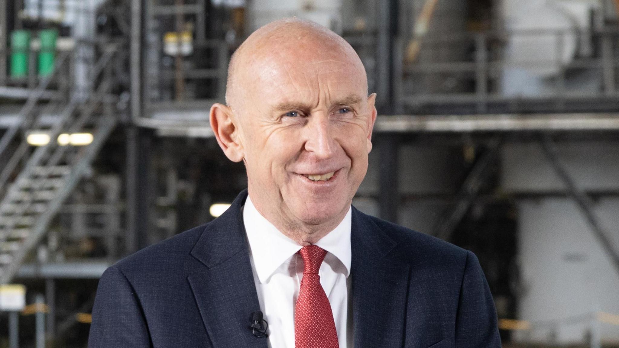 Chancellor of the Exchequer, John Healey, smiling at something off camera while dressed in a navy suit and red tie in front of oil tanks.