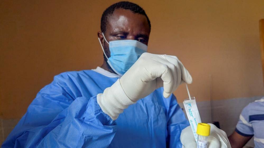 A man in surgical gloves, facemask and blue PPE put a testing stick into a test tube.