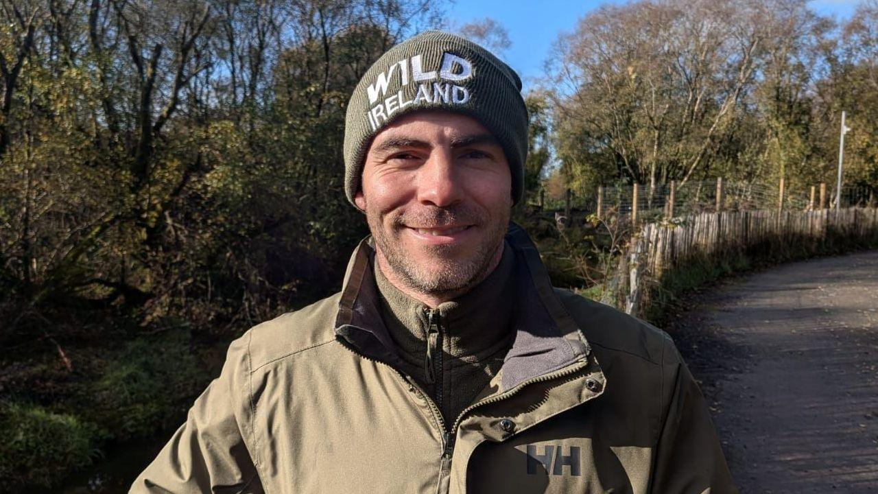 Killian McLaughlin is wearing a green coat and a woolly hat that says 'Wild Ireland'. He is smiling near to a wooden fence and grassy enclosure.