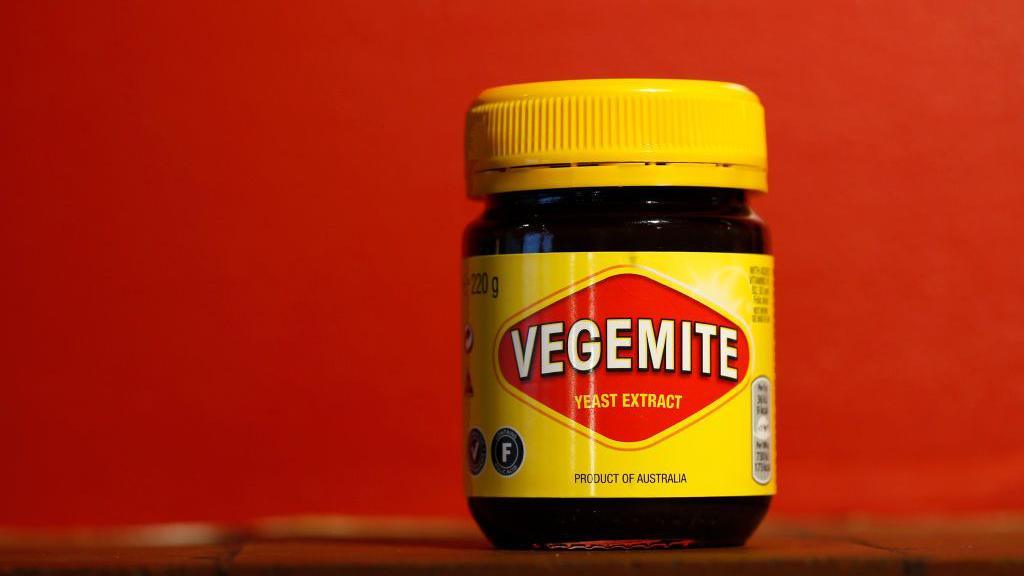 A yellow jar of Vegemite on a shelf, with a red background
