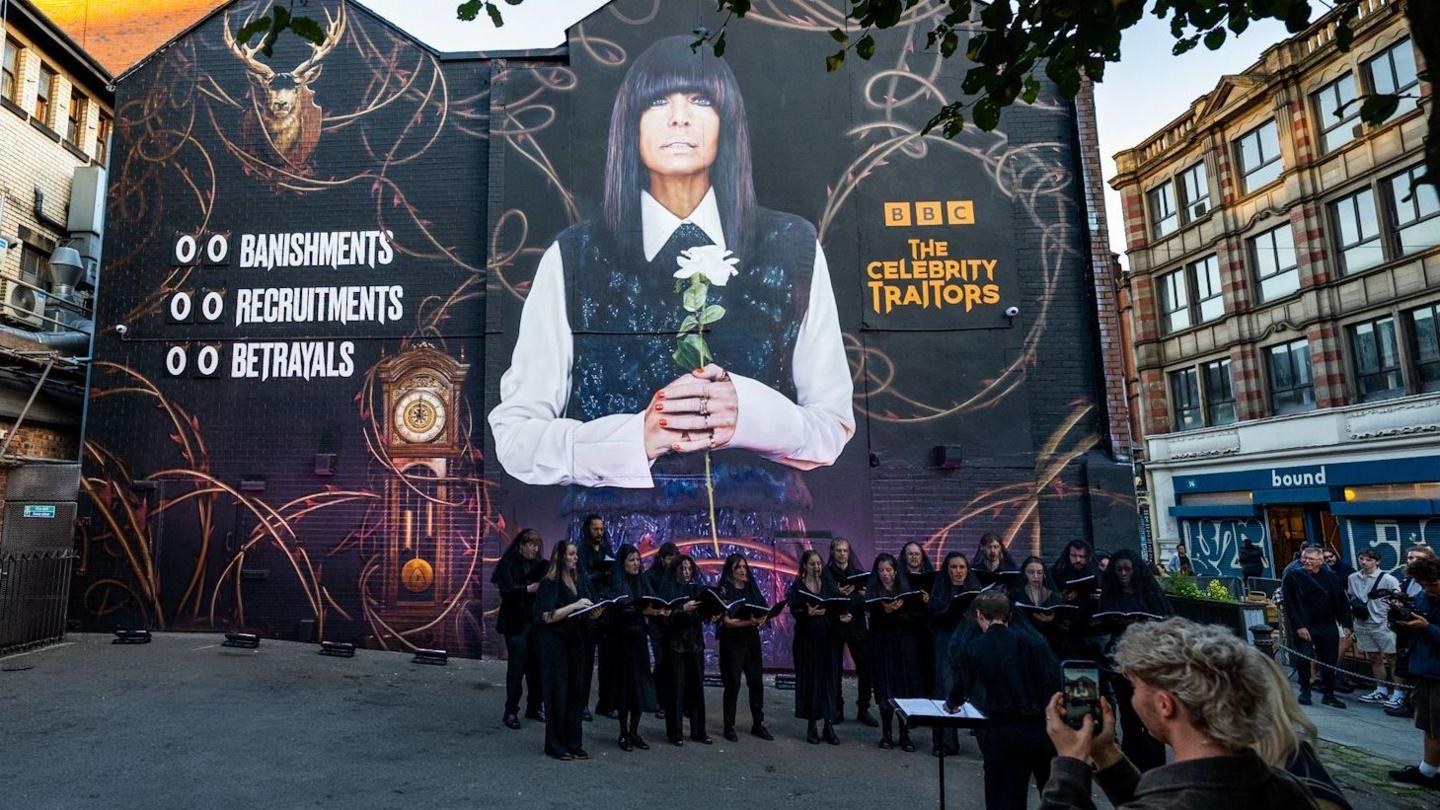 A mural featuring a stag and Claudia Winkleman. She holds a white flower in a tunic dress. There is an adjacent wall with portraits on it and a choir stands in front of the building.