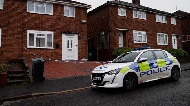 A police car is parked on the road outside a house, where police crime tape can be seen cordoning off the brick-paved driveway. The house has a white door and a white-framed front lower window.