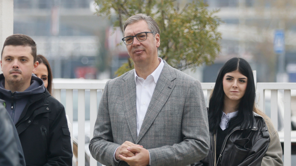 Aleksandar Vucic in a grey suit and white shirts waits in a  polling station queue