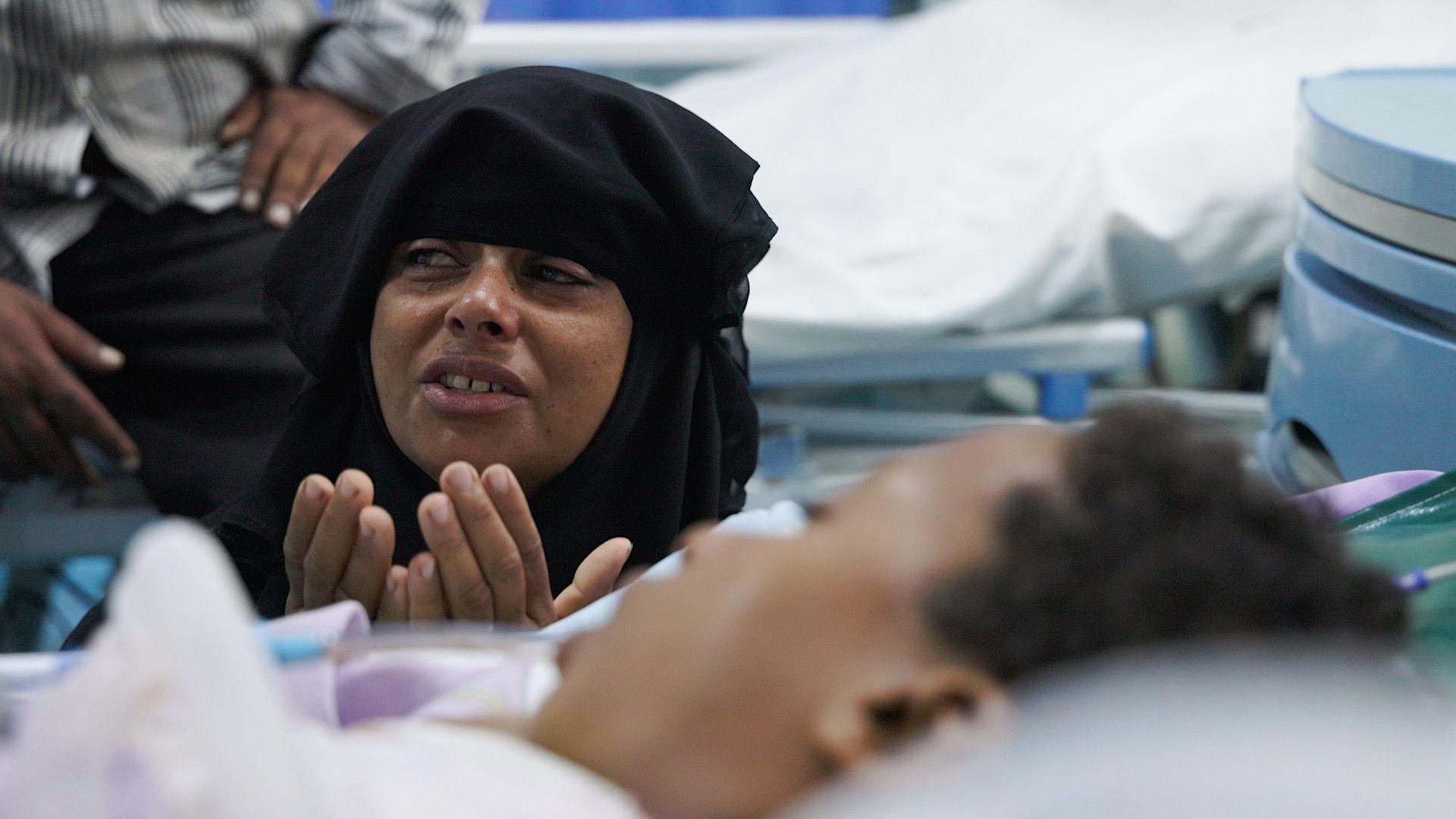 A distressed woman in a black headscarf sites next to a hospital bed, her hands are raised to her chin. A boy lies in the bed in front of her.