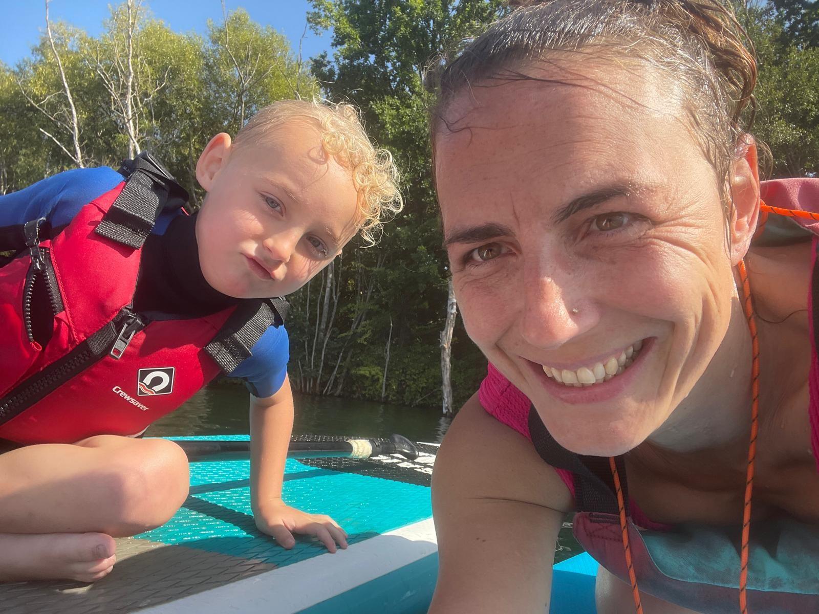 A close-up selfie of an adult and a young child on a turquoise paddleboard on a calm lake. Both are wearing life jackets, with trees and bright sunshine in the background.