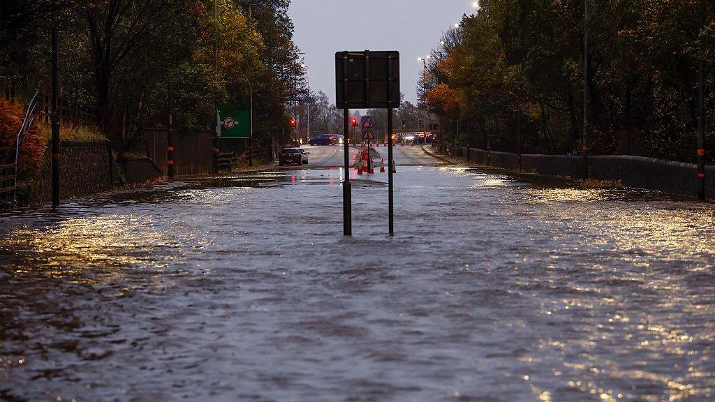 The A82 at Dumbuck Quarry is flooded, with water across the entire road. In the distance, there are a few cars on the road ahead.