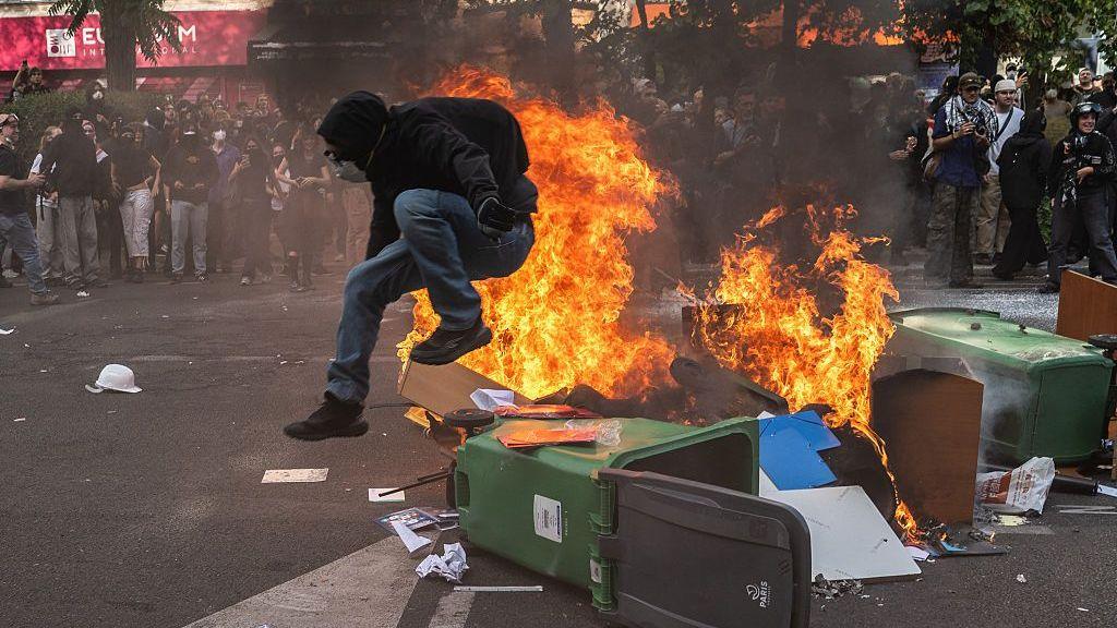A protestor in a black hoodie jumps over a wheelie bin on fire in Paris, France.