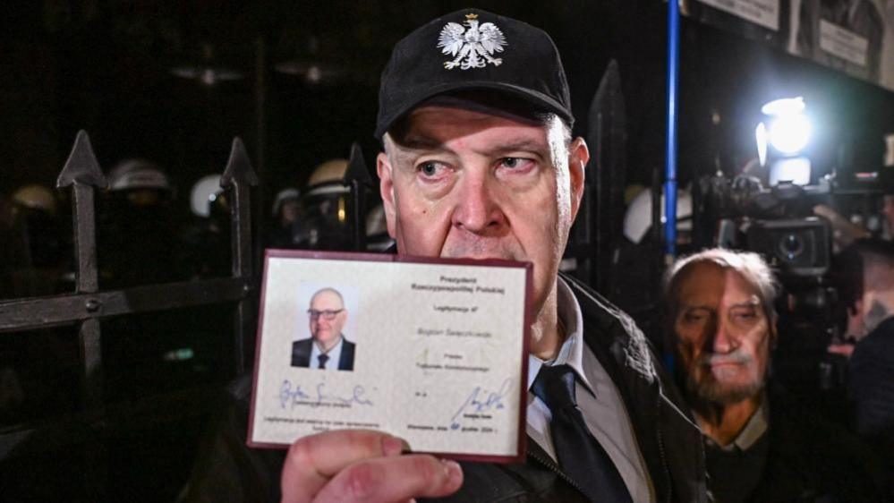 Bogdan Święczkowski holds an ID with his picture on in front of a metal fence. He is wearing a black baseball cap, with a white eagle symbol. Other people, helmets and flashing cameras can be seen in the nighttime shot.