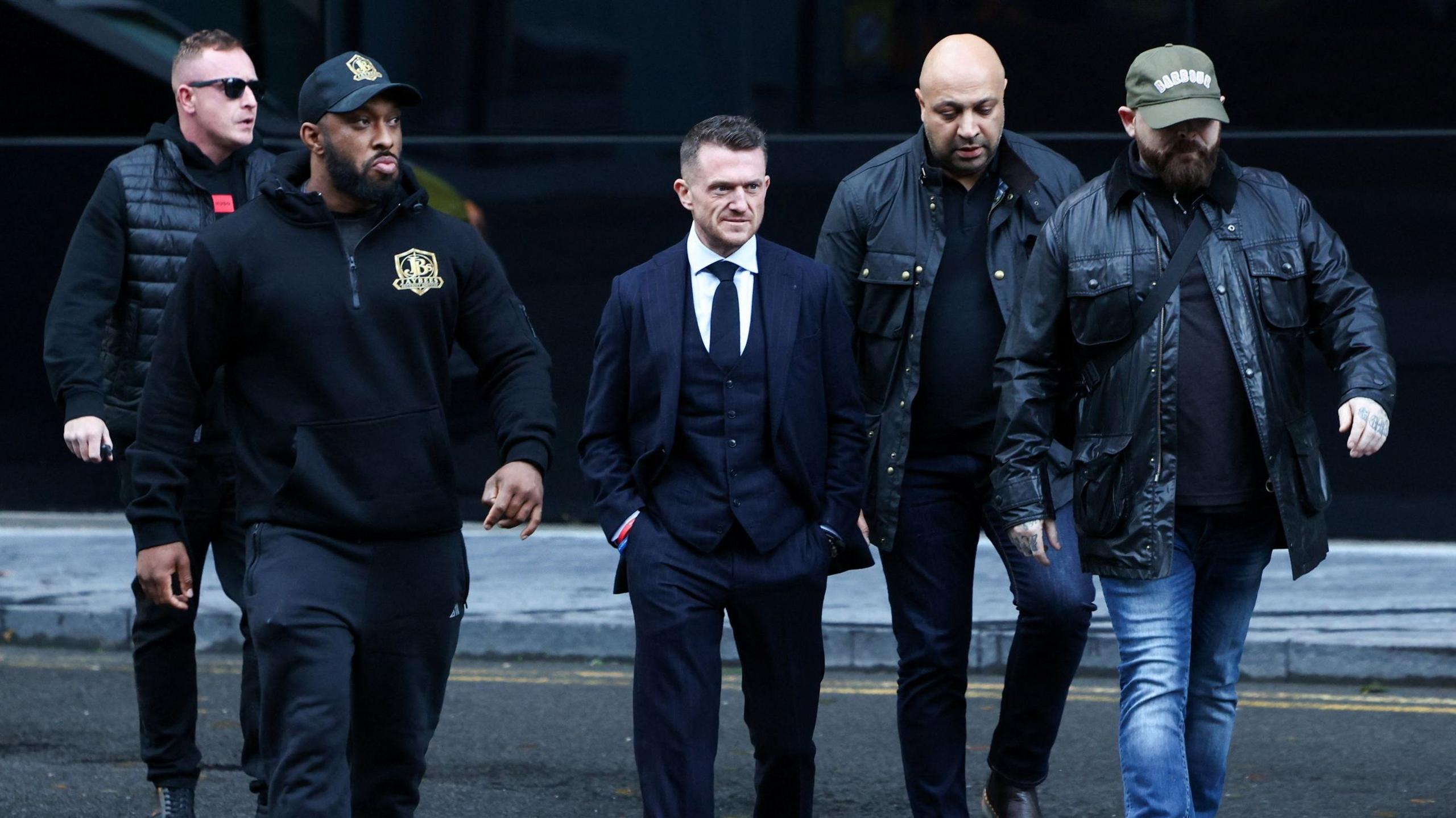 Tommy Robinson walks with his hands in his pockets, surrounded by security staff