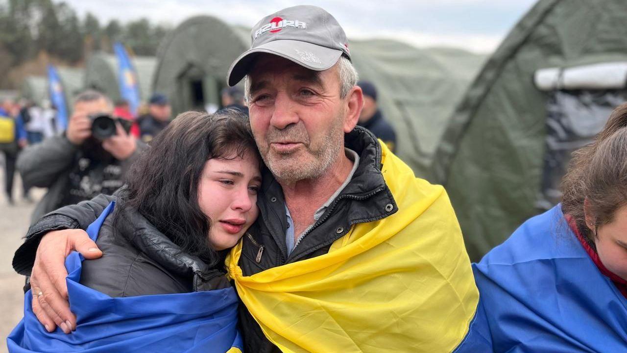 Mykhailo, wearing a grey crap holds an arm around one of his tearful daughters, whose head is placed on his chest. They are both wrapped in Ukrainian flags