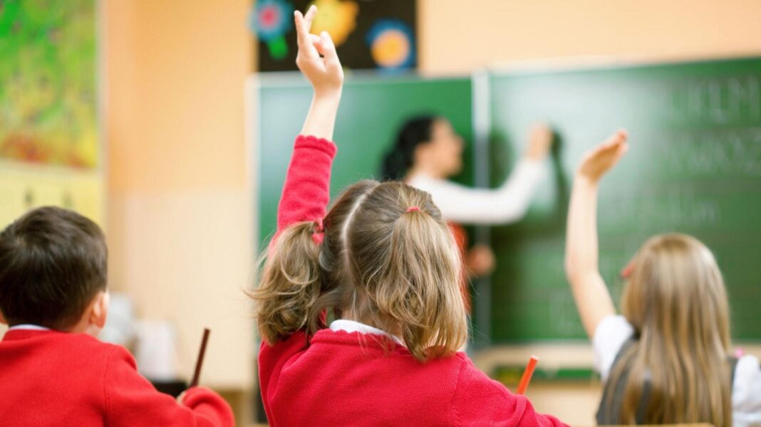 ‘Wear more clothes if it’s cold,’ schools in Northern Ireland told as energy bills rise Three children sitting at their school desks. They have their hands raised and are facing away from the camera.
