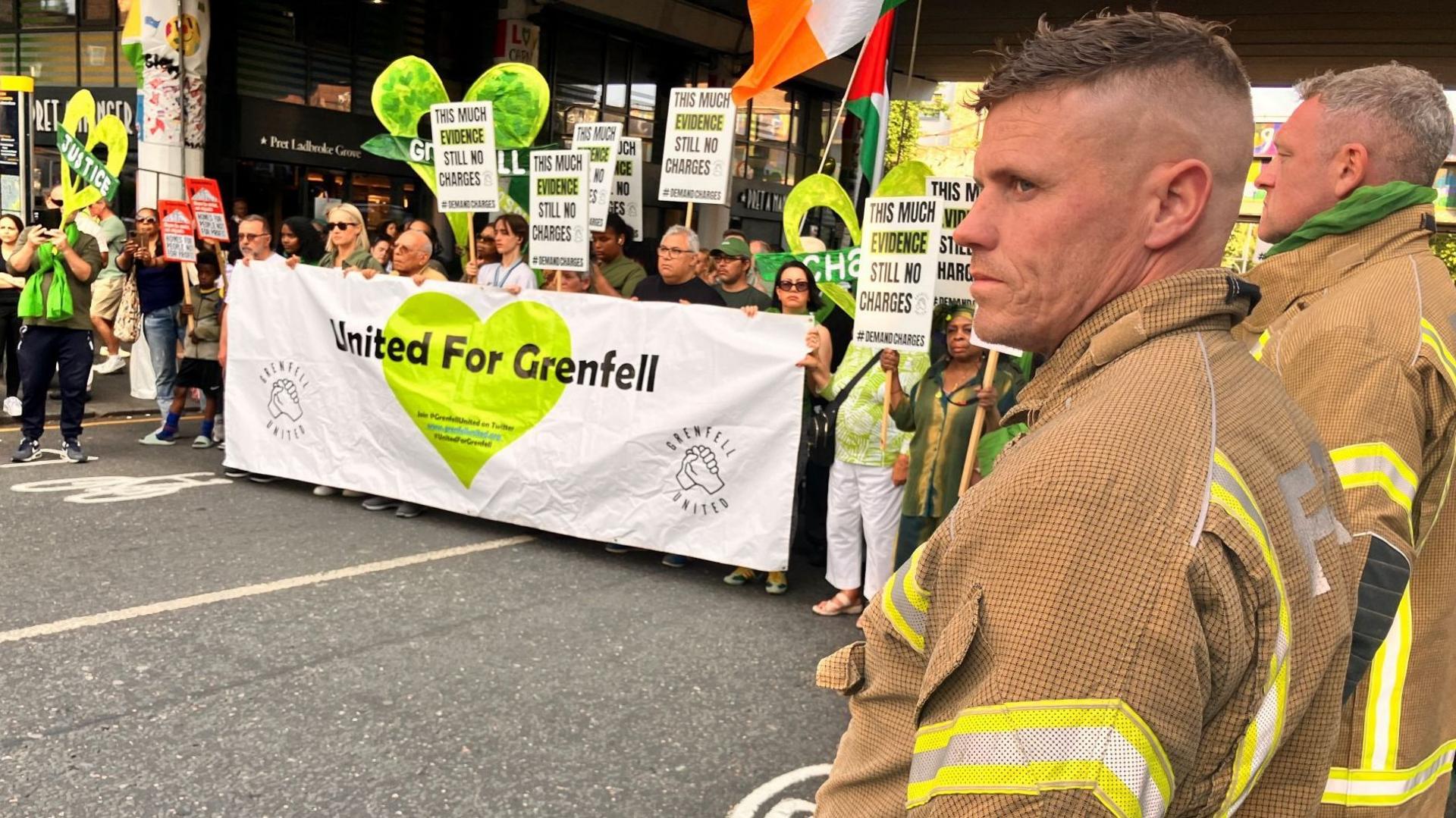 A group of people holding up signs, with messages about justice for Grenfell survivors. In the foreground, two firefighters can be seen standing with them.