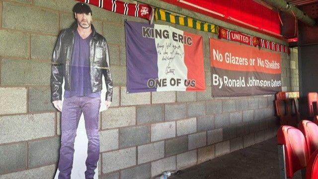 A photo showing a cardboard cut-out of former Manchester United player Eric Cantona, next to a French flag with the words 'King Eric one of us' written on it, leaning against a wall at the back of a stand at Broadhurst Park.